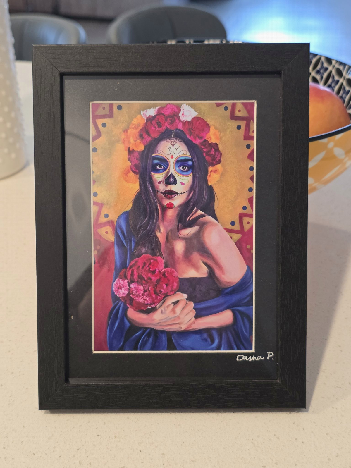 Catrina signed matted framed art print by Dasha Prudnikova, a colorful Day of the Dead portrait holding red flowers in a black frame with black mat.