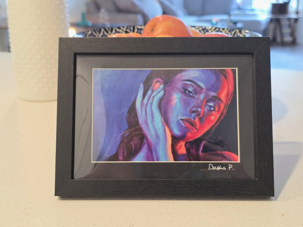 Still Between Thoughts signed matted framed art print by Dasha Prudnikova, a neon blue and pink portrait of a woman with her hand on her face in a black frame with black mat.