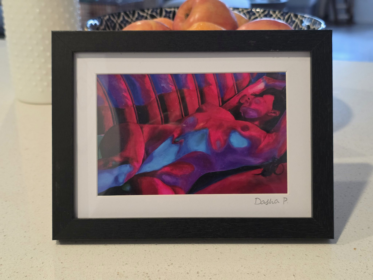 Unwind signed matted framed art print by Dasha Prudnikova, a reclining figure in vivid red and blue tones in a black frame with white mat.